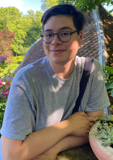 Image Description: Moises is a Hispanic male with brown hair with a side part. He is wearing a gray shirt with a bag around his right shoulder. There are flowers in the background and he is leaning against a brick wall.